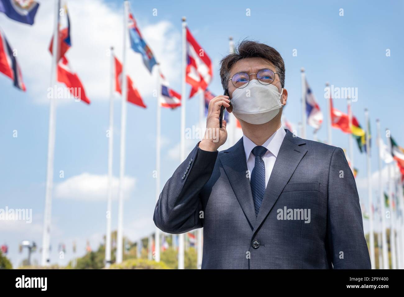 Homme d'affaires asiatique d'âge moyen portant un masque et utilisant un smartphone sous différents drapeaux nationaux qui flottent dans le vent. Banque D'Images