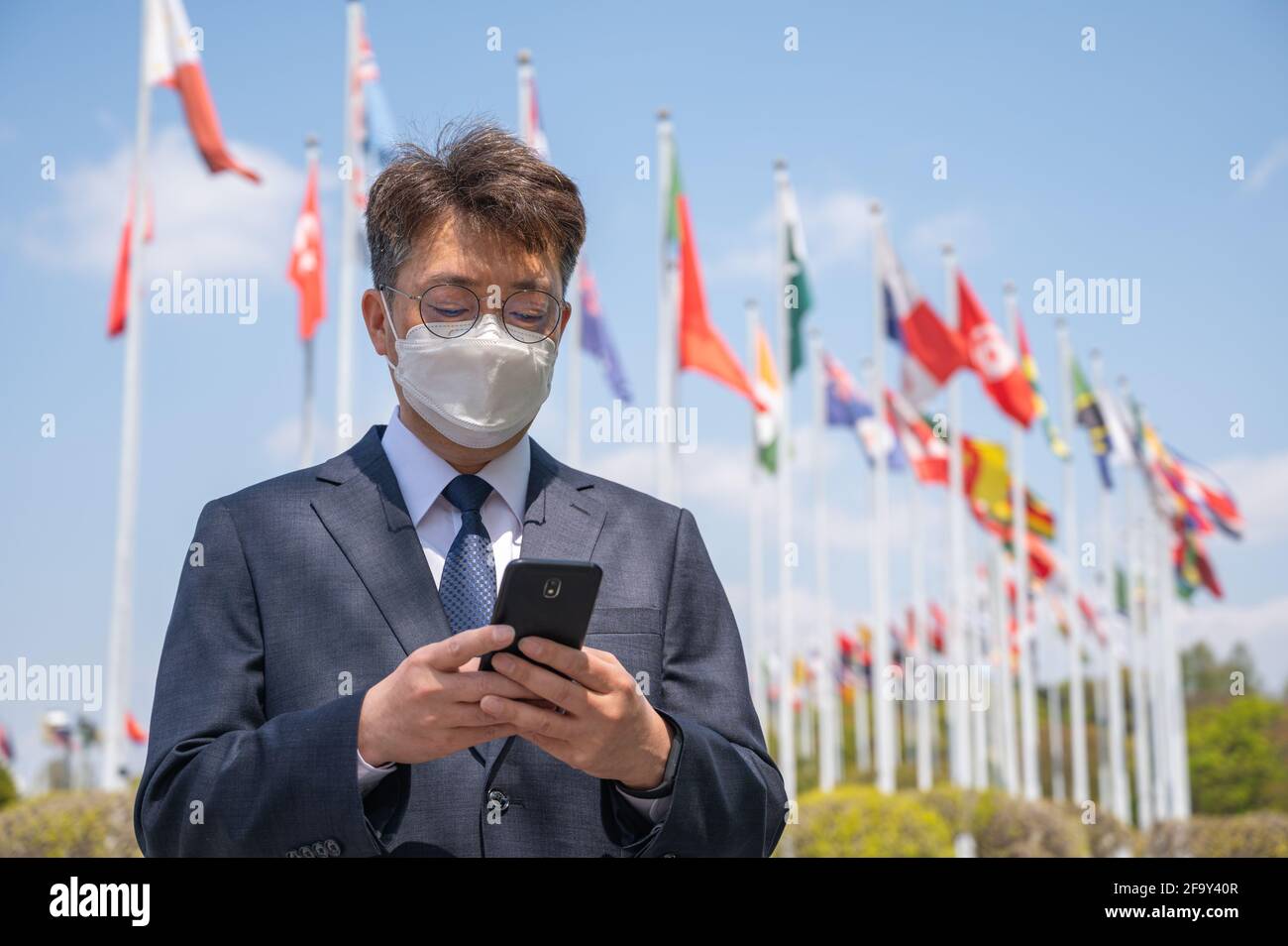 Homme d'affaires asiatique d'âge moyen portant un masque et utilisant un smartphone sous différents drapeaux nationaux qui flottent dans le vent. Banque D'Images
