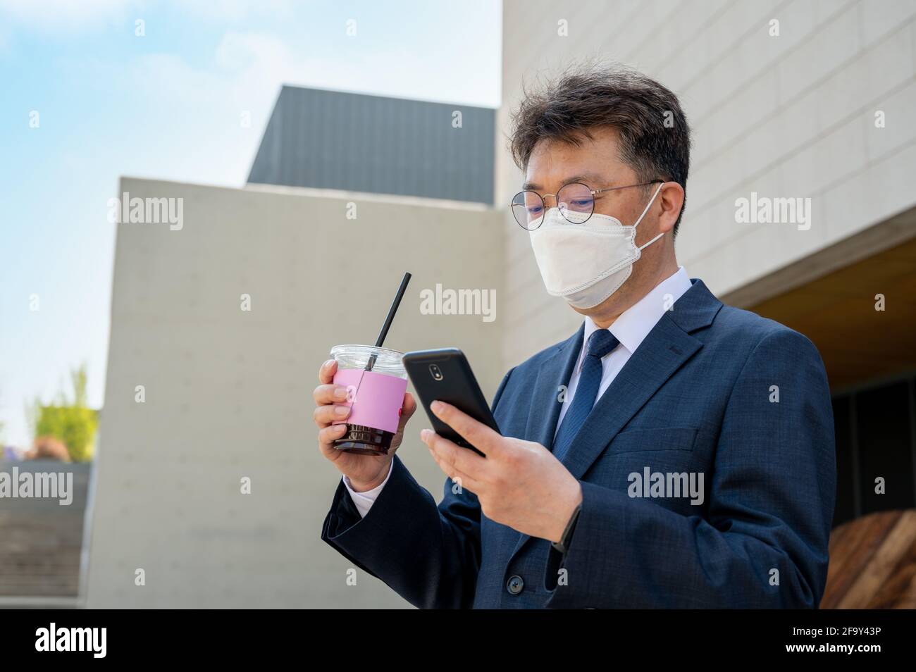 Un homme d'affaires asiatique d'âge moyen dans la ville portant un masque, tenant une tasse de café et utilisant un smartphone. Banque D'Images