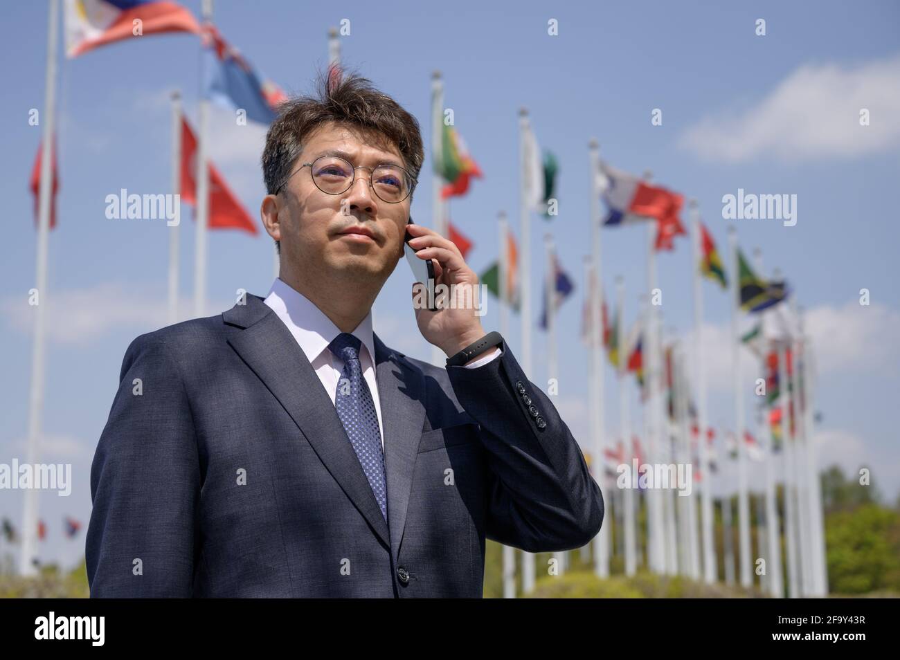 Homme d'affaires asiatique d'âge moyen utilisant un smartphone sous différents drapeaux nationaux qui flottent dans le vent. Banque D'Images