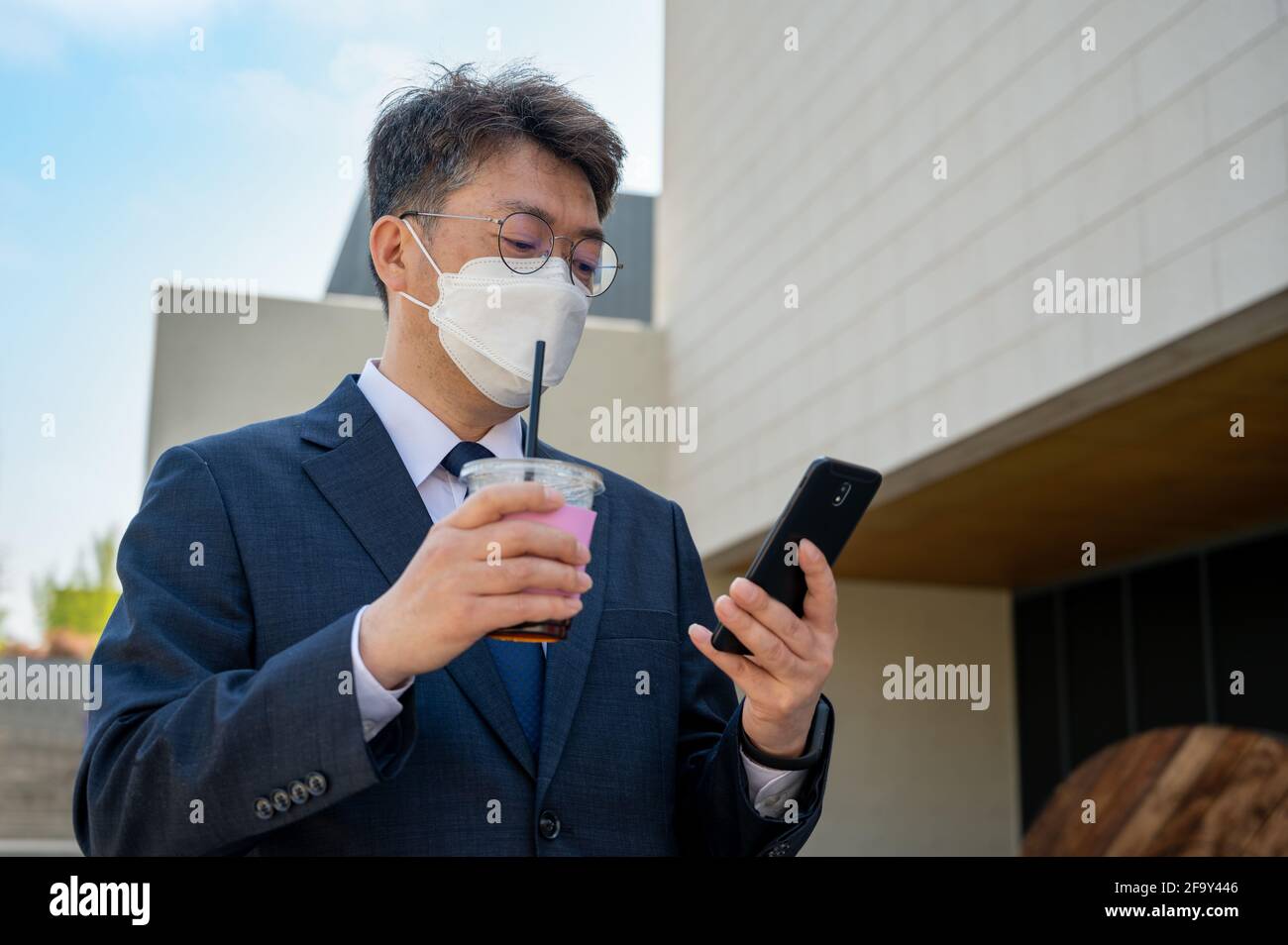 Un homme d'affaires asiatique d'âge moyen dans la ville portant un masque, tenant une tasse de café et utilisant un smartphone. Banque D'Images