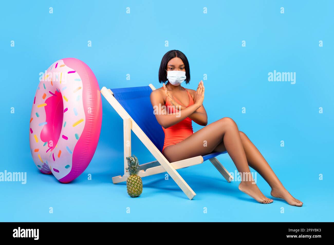 Interdiction de voyager du coronavirus. Femme noire en maillot de bain et masque de visage montrant UN GESTE D'ARRÊT tout en étant assise dans la chaise longue Banque D'Images