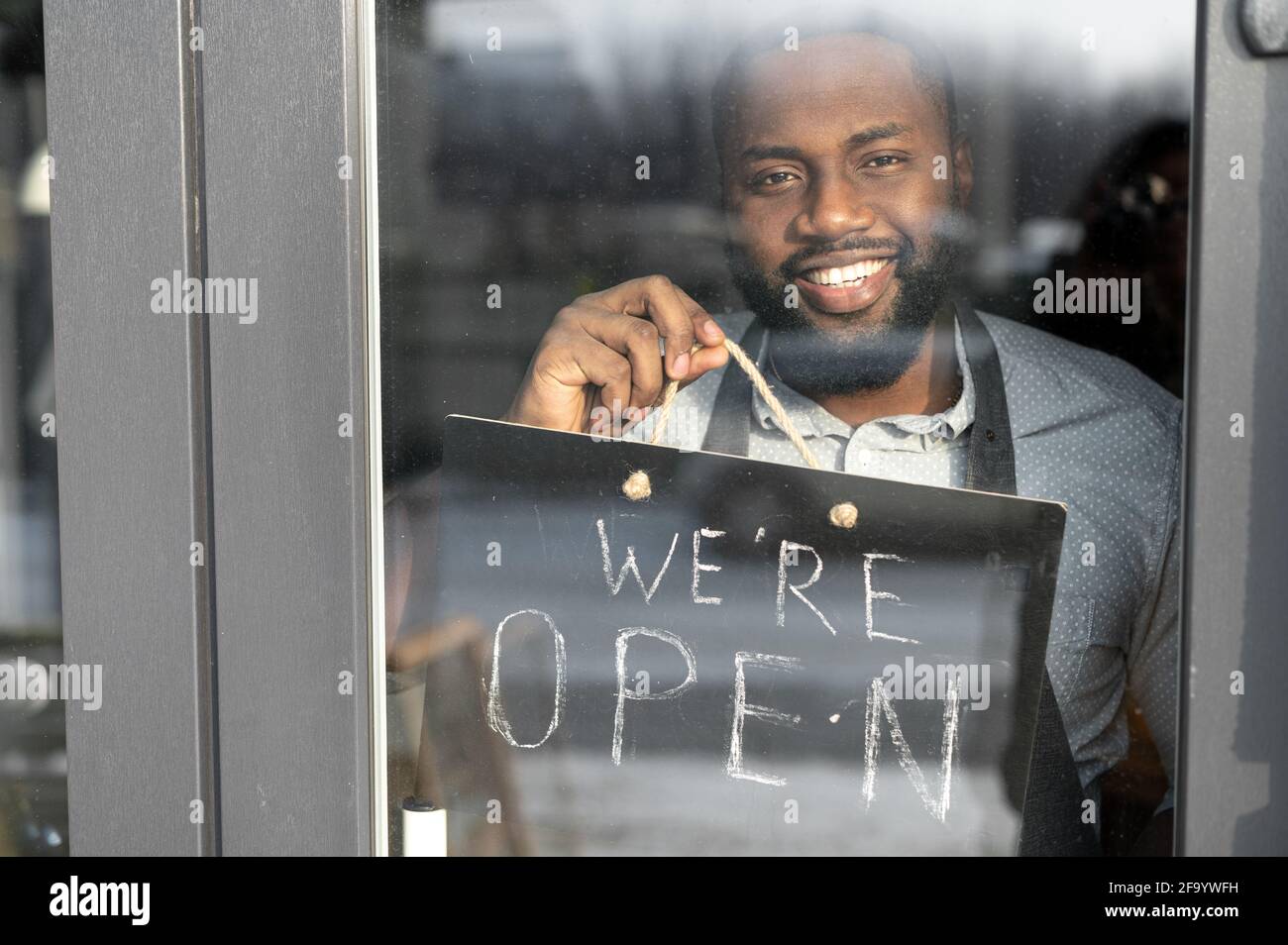 Vue à travers la porte en verre sur un homme africain de serveur Manager avec un panneau ouvert dans sa main. Un propriétaire de café multiracial vous invite à visiter le lieu de réouverture Banque D'Images