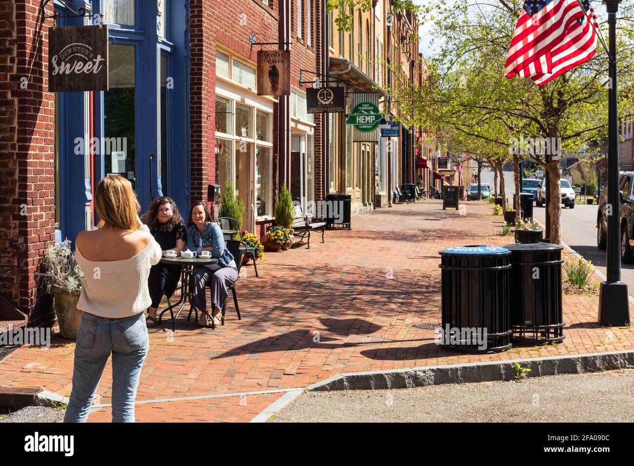 JONESBOROUGH, TN, USA--9 AVRIL 2021:une femme mince et blonde avec dos à la caméra prend une photo de deux amis assis à une table de trottoir. Banque D'Images