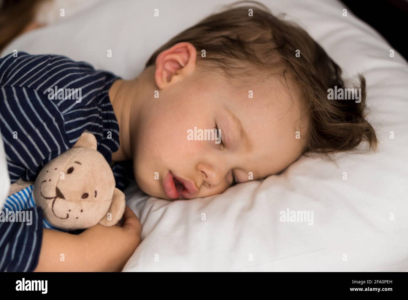 Portrait authentique malade mignon petit petit garçon d'âge préscolaire caucasien dans le sommeil bleu avec l'ours en peluche sur le lit blanc. Enfant reposant à l'heure du déjeuner. Soins Banque D'Images