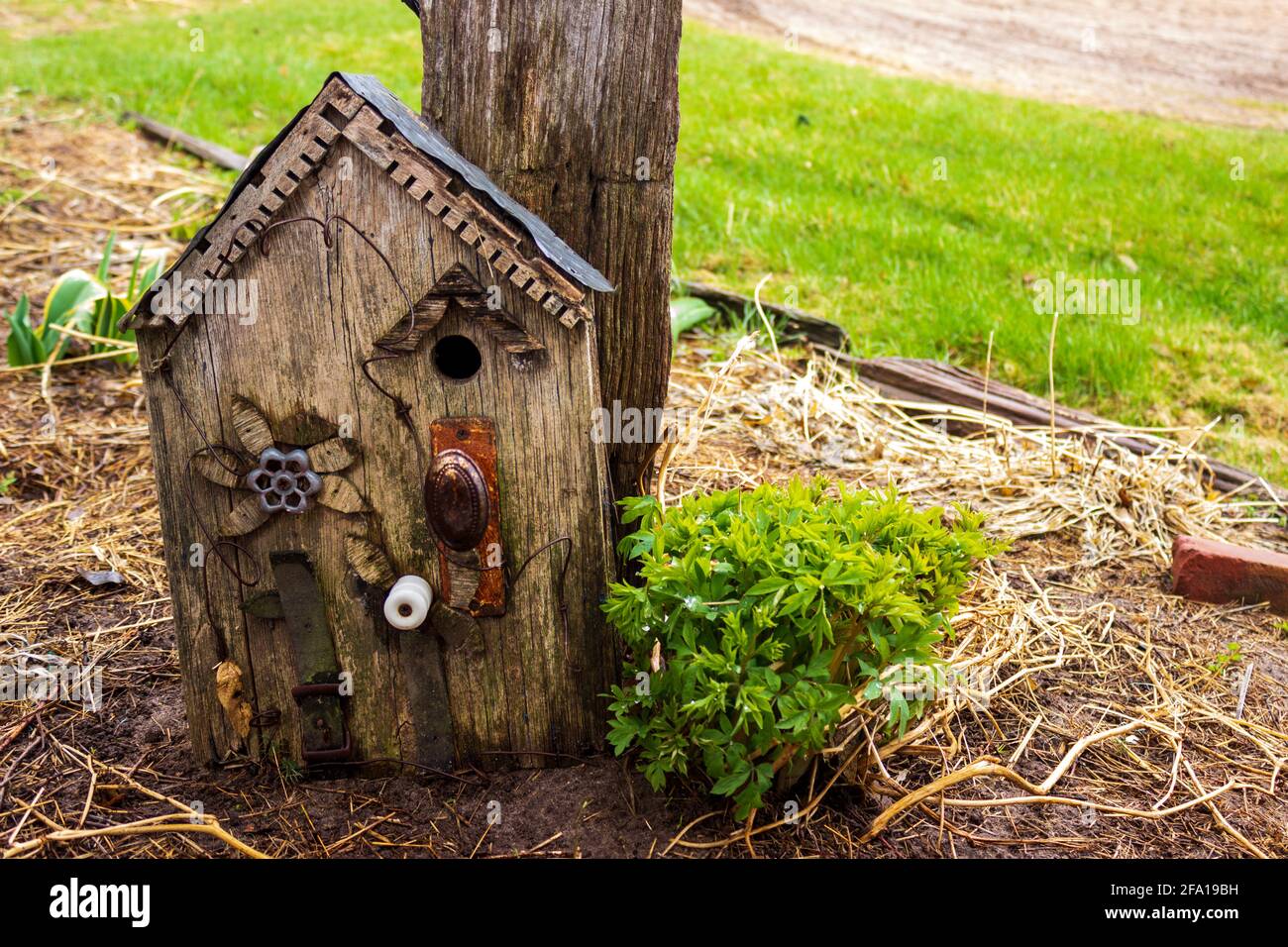 Un birdhouse rustique fait d'une poignée de porte réutilisée, poignée de robinet d'eau, ceinture, isolant, étain et vieux bois décoratif penche contre un poteau. Banque D'Images