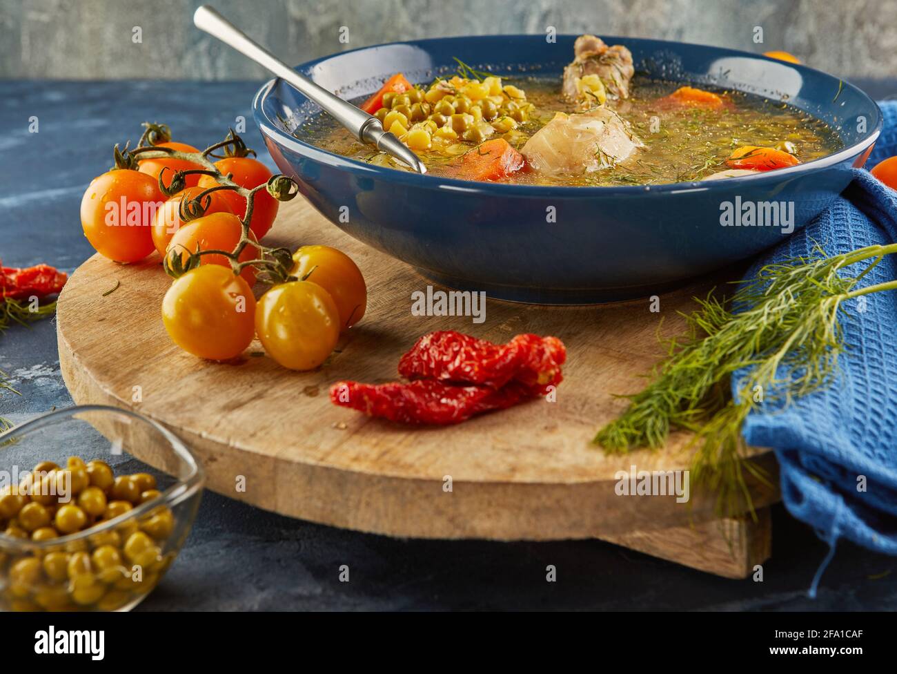 Soupe aux pois avec poulet, carottes, tomates et herbes dans une assiette profonde sur bois avec serviette sur fond bleu. Banque D'Images