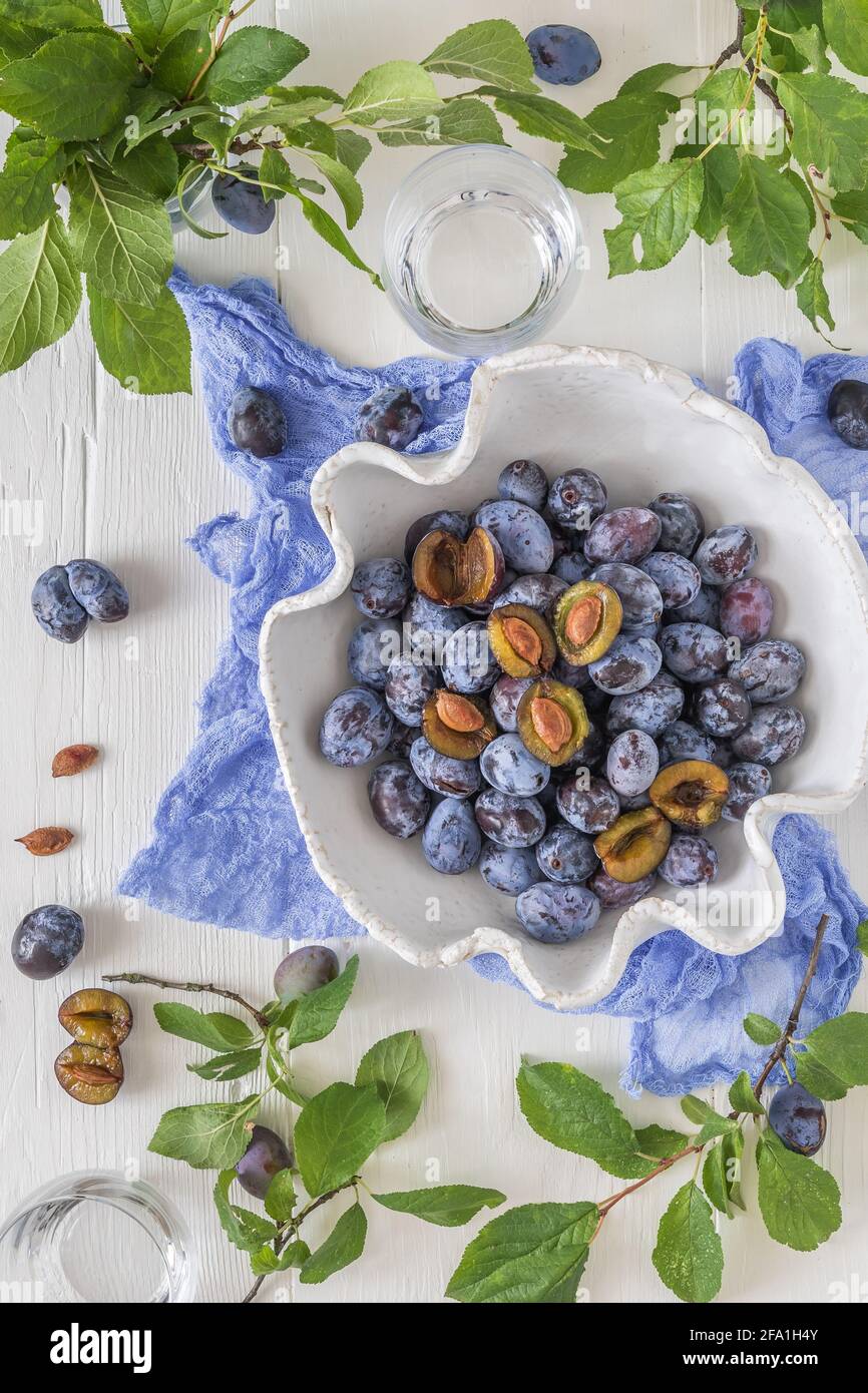 Bol blanc incurvé rempli de prunes mûres, dont certaines sont tranchées, sur une table en bois blanc avec des feuilles vertes. Vue de dessus. Banque D'Images