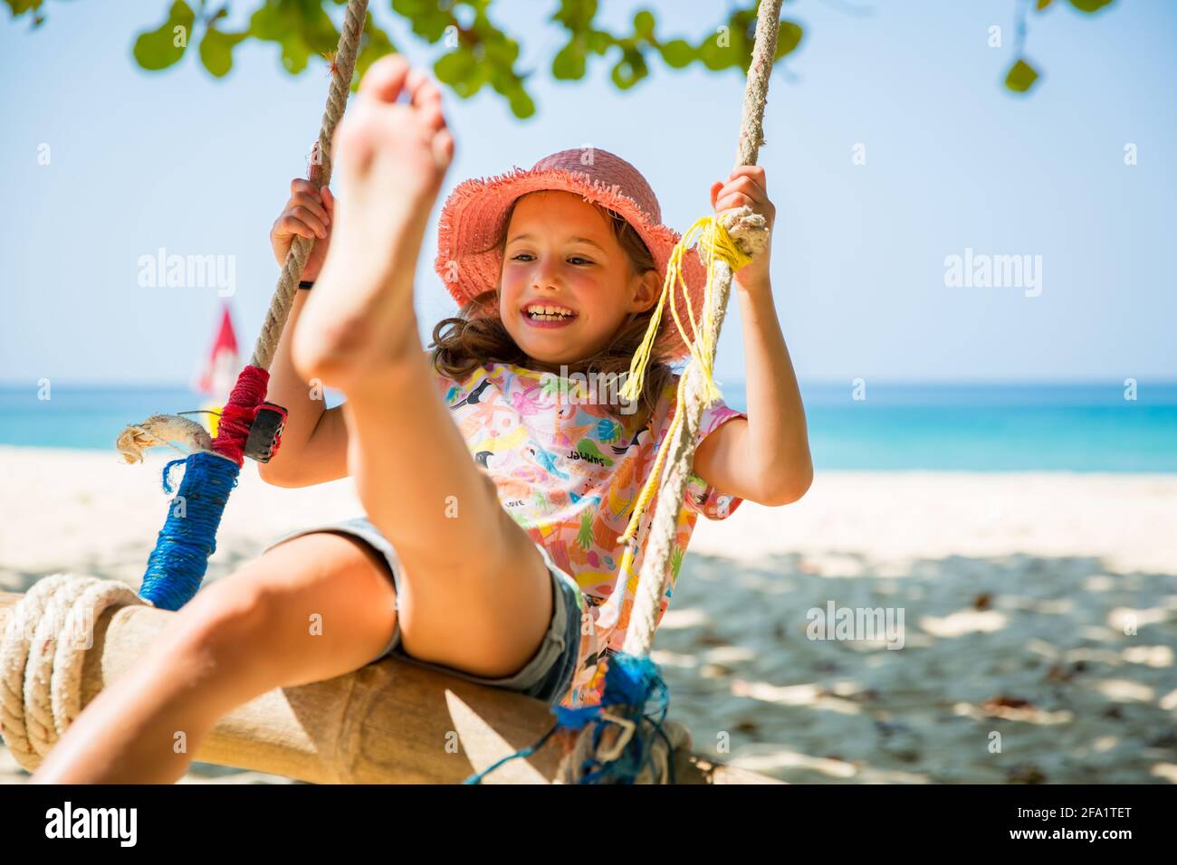 Bonne mignonne petite fille riant et balançant sur l'arbre à la plage. Belle journée ensoleillée d'été, mer turquoise, rochers, sable blanc, Banque D'Images