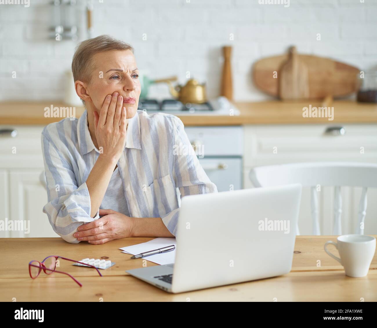 Une femme mûre frustrée touchant sa joue, souffrant de maux de dents tout en étant assise dans la cuisine à la maison tout en travaillant à la maison sur un ordinateur portable, une attention sélective sur la femme. Concept de santé dentaire Banque D'Images