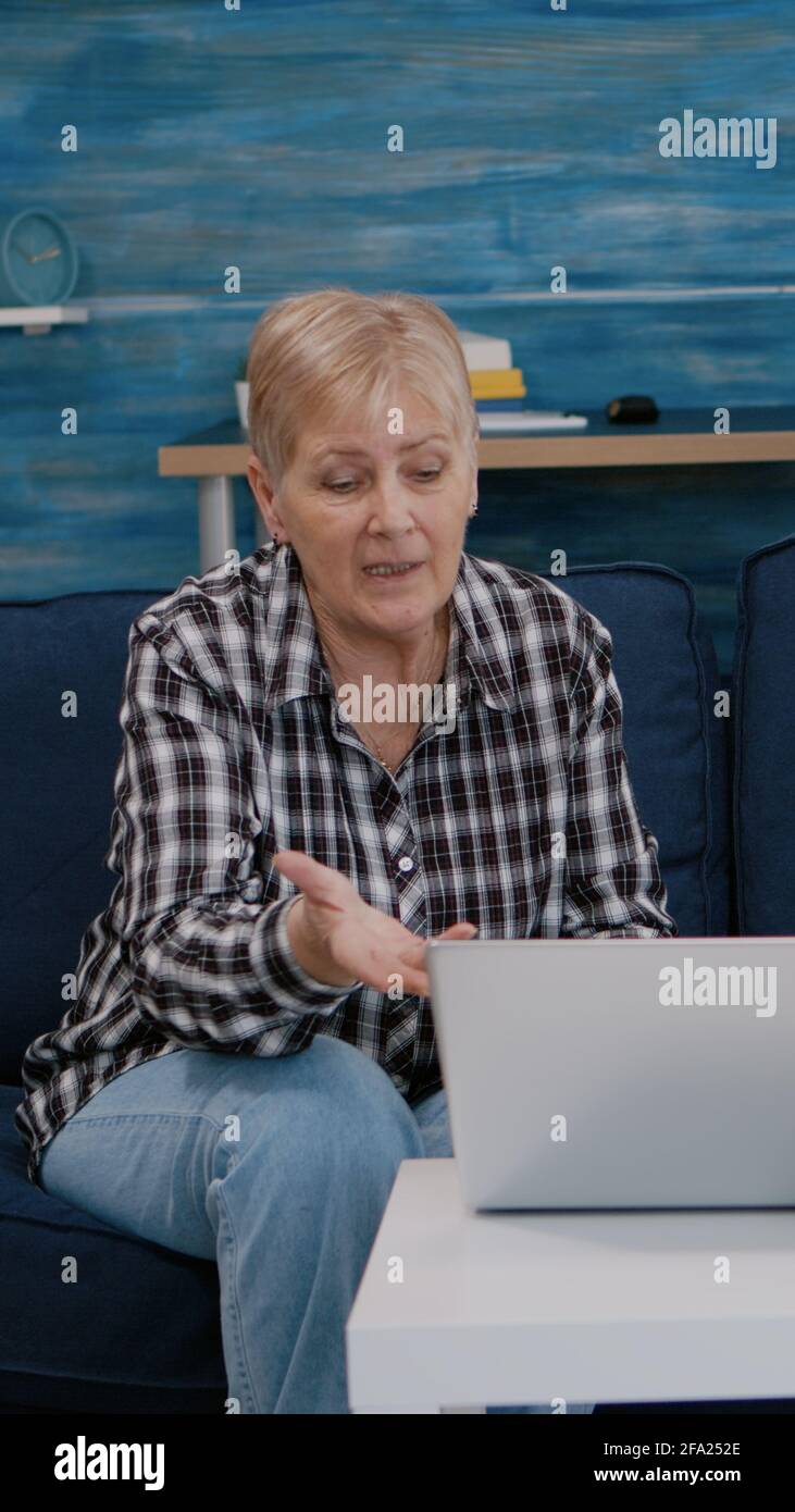 Un couple expérimenté souriant et enthousiaste regardant un ordinateur portable se faire passer pendant que le magnétoscope est assis sur un canapé. Un mari et une femme à la retraite souriant lors d'une réunion virtuelle se reposant sur un canapé dans une salle confortable. Banque D'Images