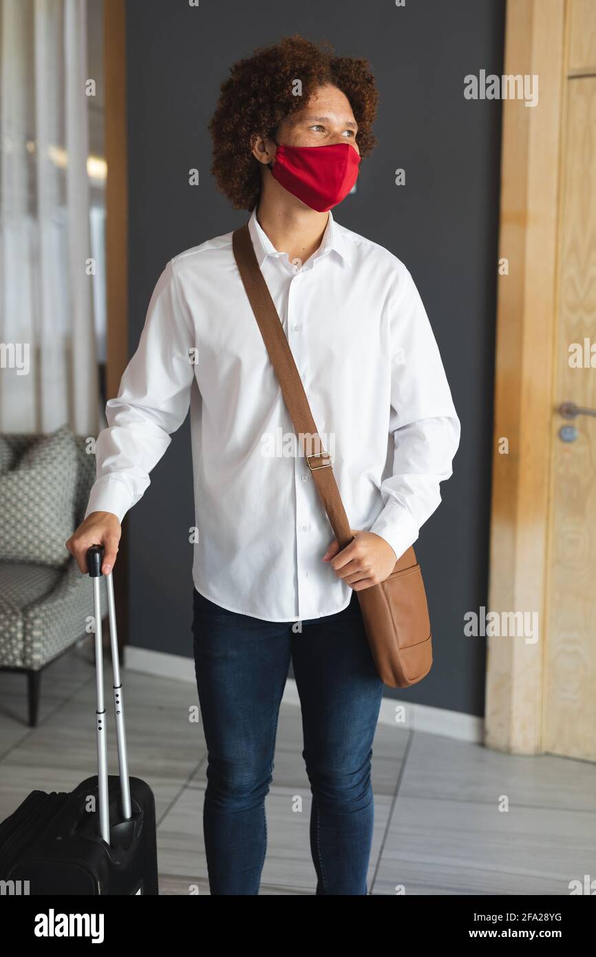 Portrait d'un homme de race mixte portant un masque de visage debout hall de l'hôtel avec sac à bandoulière et valise Banque D'Images