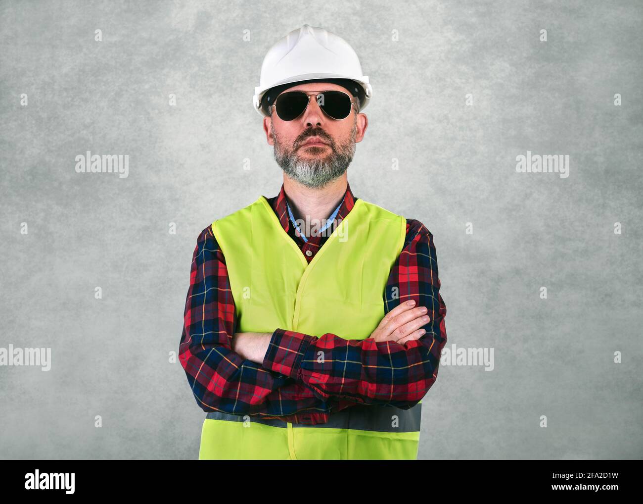 Homme ouvrier de construction avec un casque blanc et des lunettes de soleil avec bras croisés et gilet de sécurité debout sur fond gris Banque D'Images