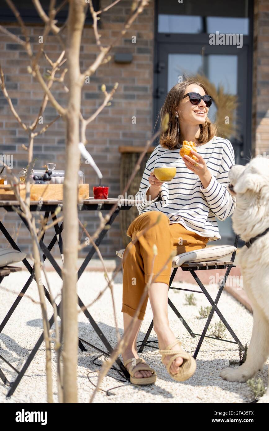 Femme gaie assise à une table dans le jardin près de la maison avec un grand chien blanc et déjeuner. Passez du temps à l'extérieur. Banque D'Images