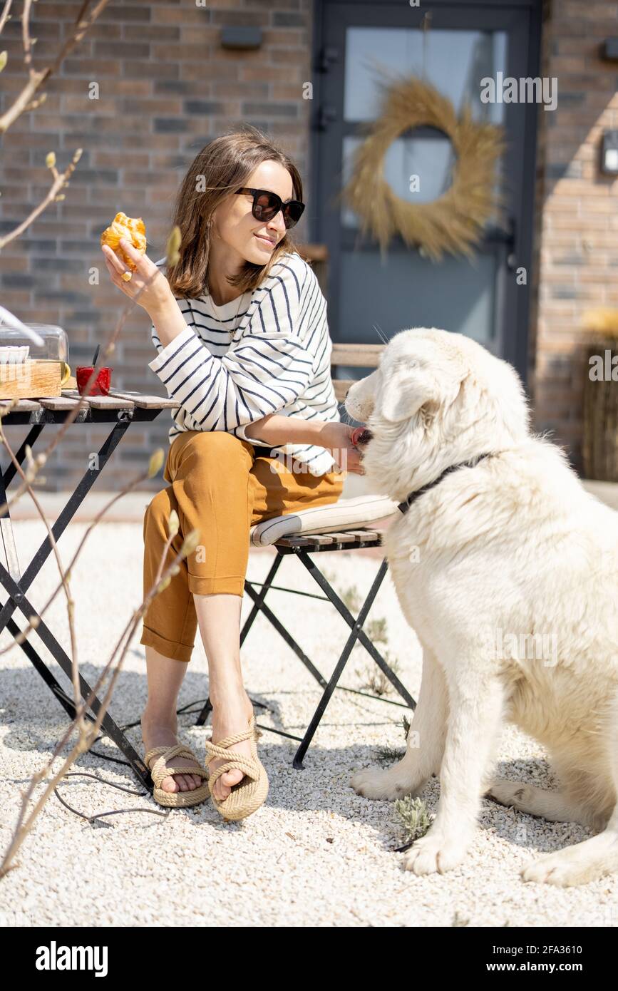 Femme gaie assise à une table dans le jardin près de la maison avec un grand chien blanc et déjeuner. Passez du temps à l'extérieur. Banque D'Images