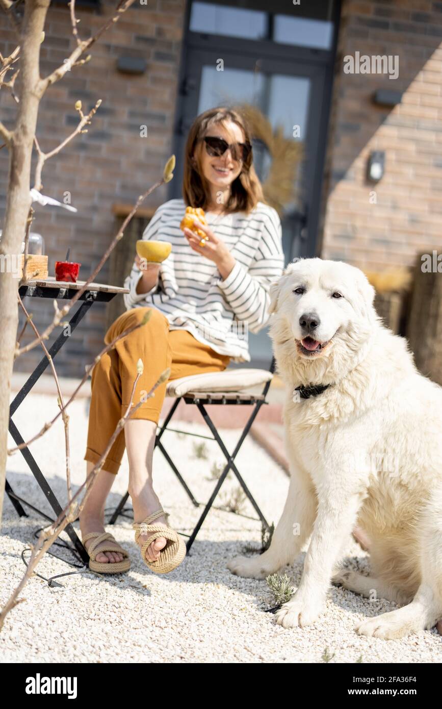 Femme gaie assise à une table dans le jardin près de la maison avec un grand chien blanc et déjeuner. Passez du temps à l'extérieur. Banque D'Images