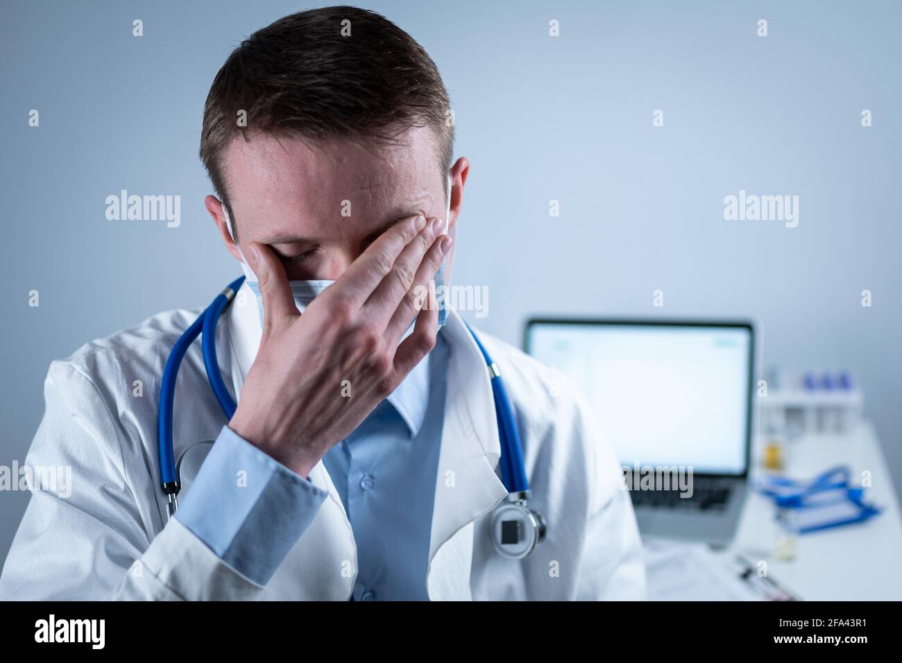 Jeune médecin européen allemand regardant l'appareil photo portant des lunettes et un masque dans le bureau de la clinique sur le lieu de travail avec le regard fatigué. Le sujet est le dur travail de Banque D'Images