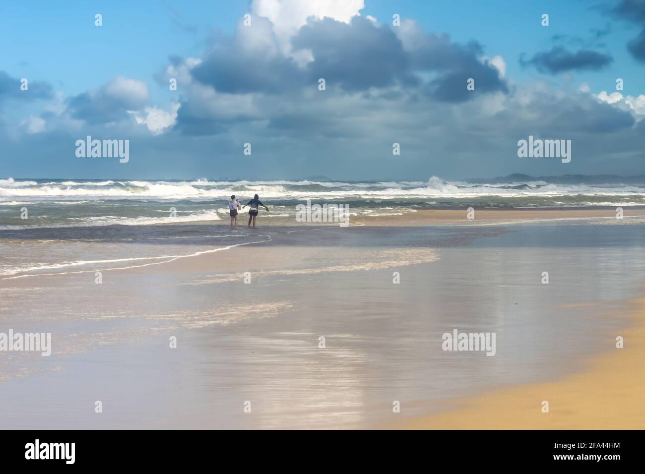 Deux femmes touristes - une avec les bras débordés aiment se tenir dehors sur la plage humide sous le ciel orageux avec l'océan turbulent dedans arrière-plan Banque D'Images