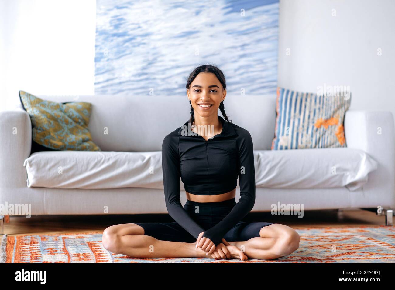 Jeune femme afro-américaine détendue dans des vêtements de sport noirs, assis sur le tapis à la maison dans la position lotus, faisant du yoga, menant un style de vie sain, regardant la caméra, souriant Banque D'Images