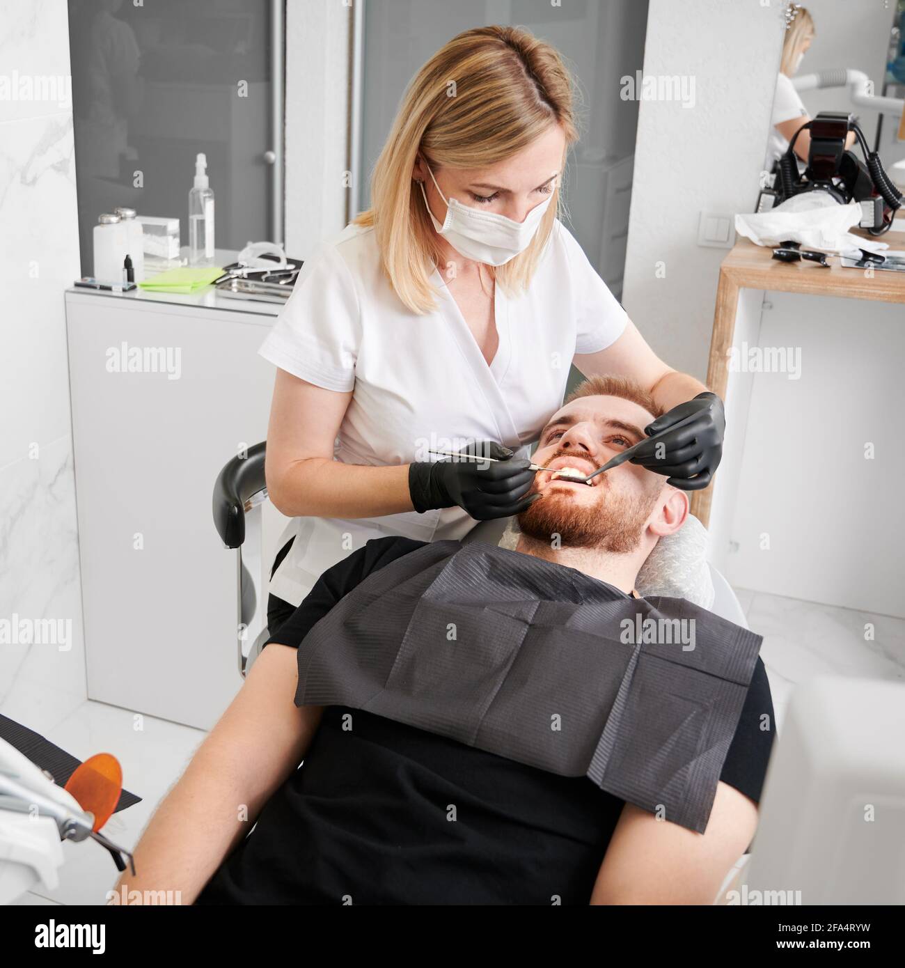 Vue du dessus du patient allongé sur une chaise de dentiste, portant un bavoir noir, une femme dentiste effectuant un contrôle des dents avec des outils dentaires, portant des gants noirs, un masque facial et un uniforme. Concept de la dentisterie Banque D'Images