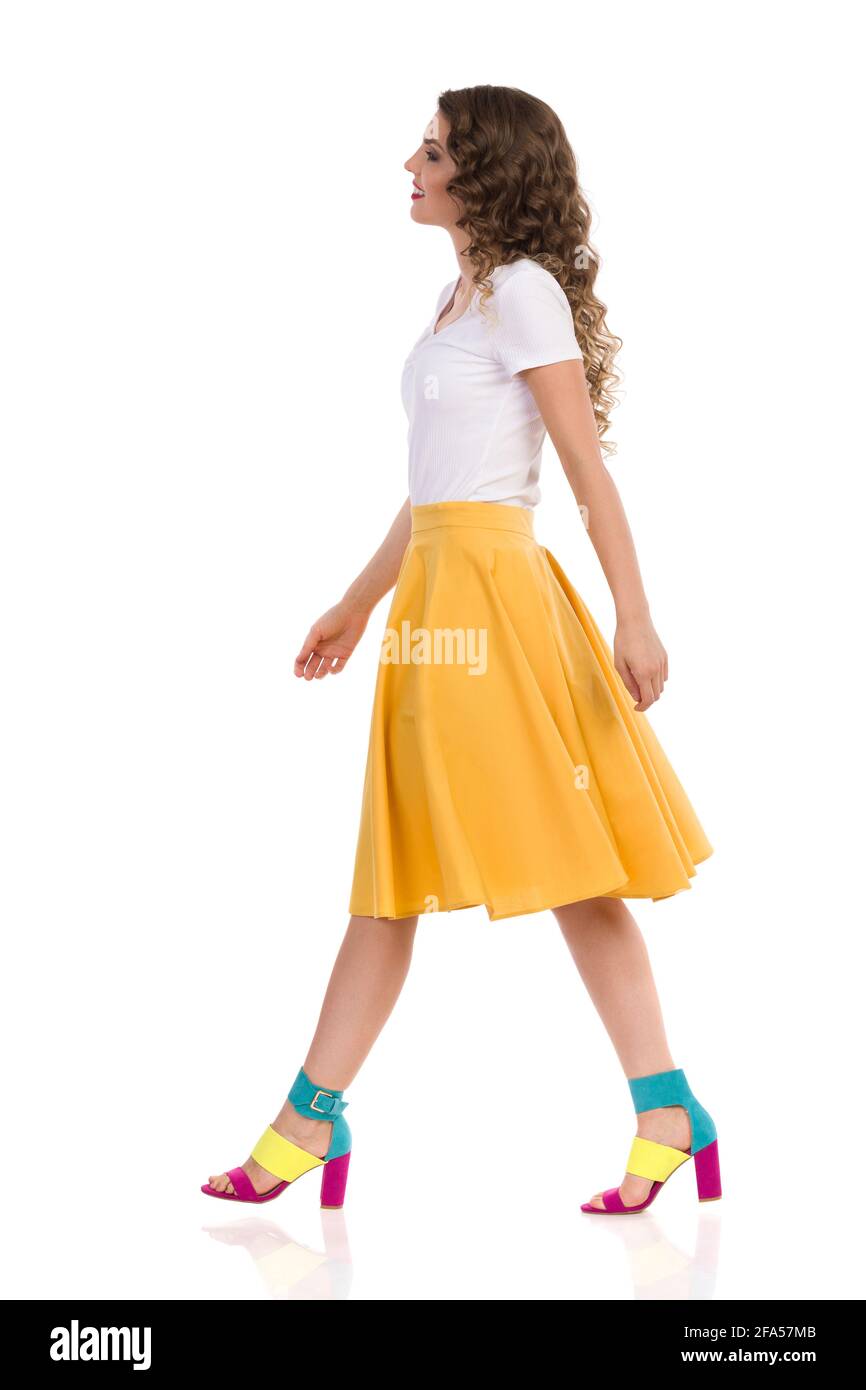 Belle jeune femme en haut blanc, jupe jaune et hauts talons hauts colorés marche et regarder loin. Vue latérale. Prise de vue en studio sur toute la longueur isolée sur W Banque D'Images