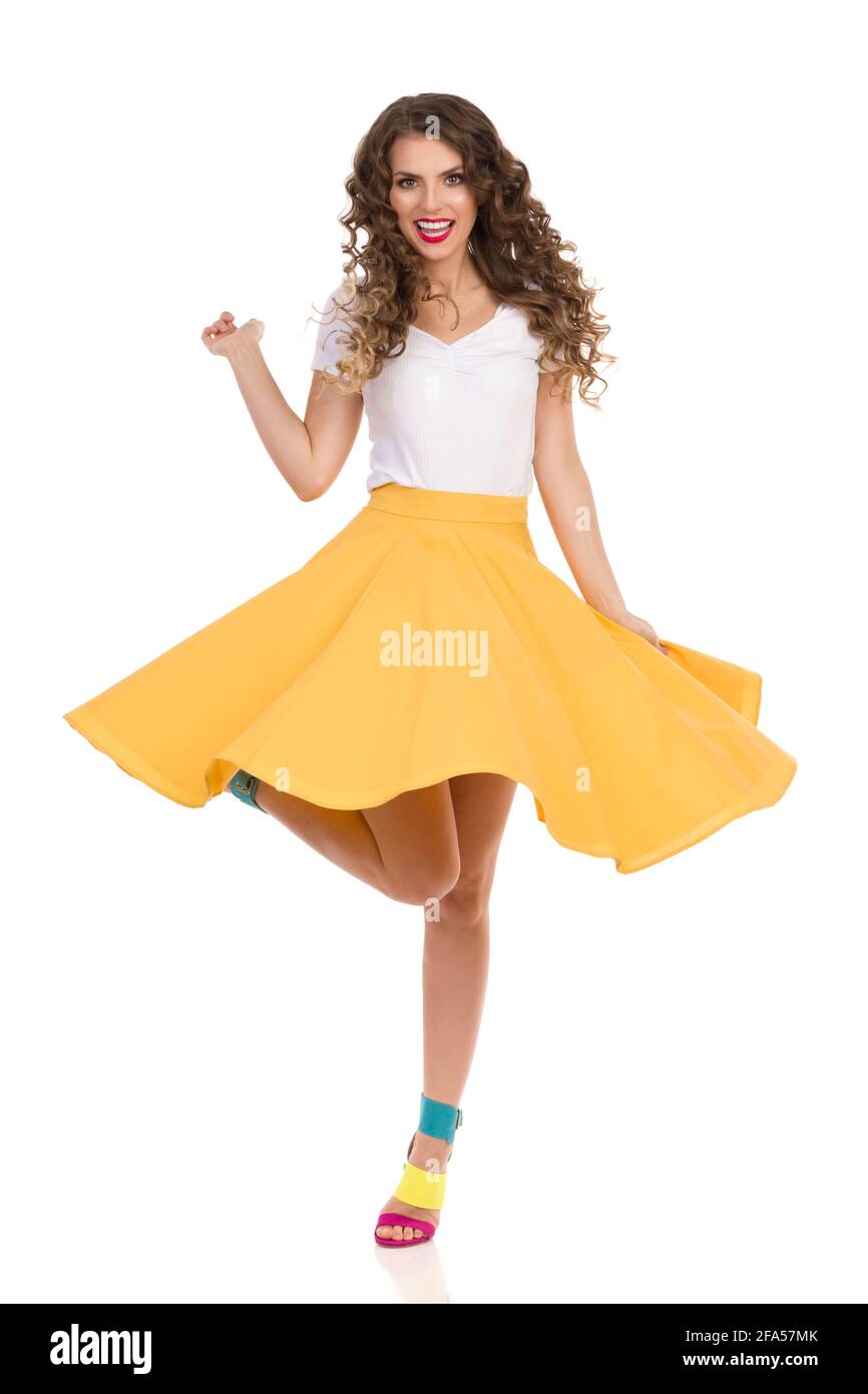 Belle jeune femme en jupe jaune, haut blanc et hauts talons hauts colorés chante sur une jambe et chante. Prise de vue en studio sur toute la longueur isolée sur blanc. Banque D'Images