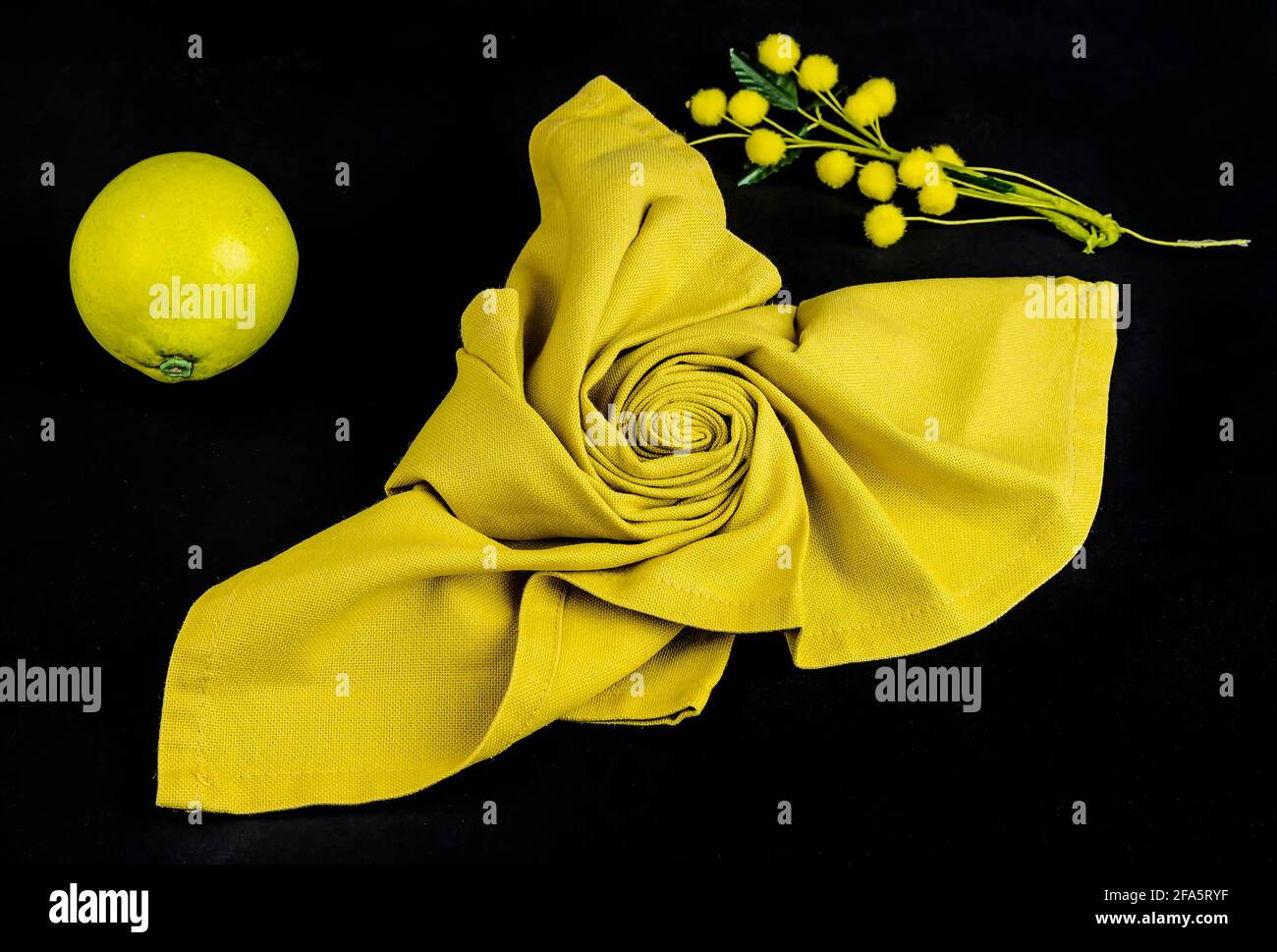 Une serviette en tissu jaune de forme florale, un citron et un bouquet de fleurs mimosa sur fond noir Banque D'Images