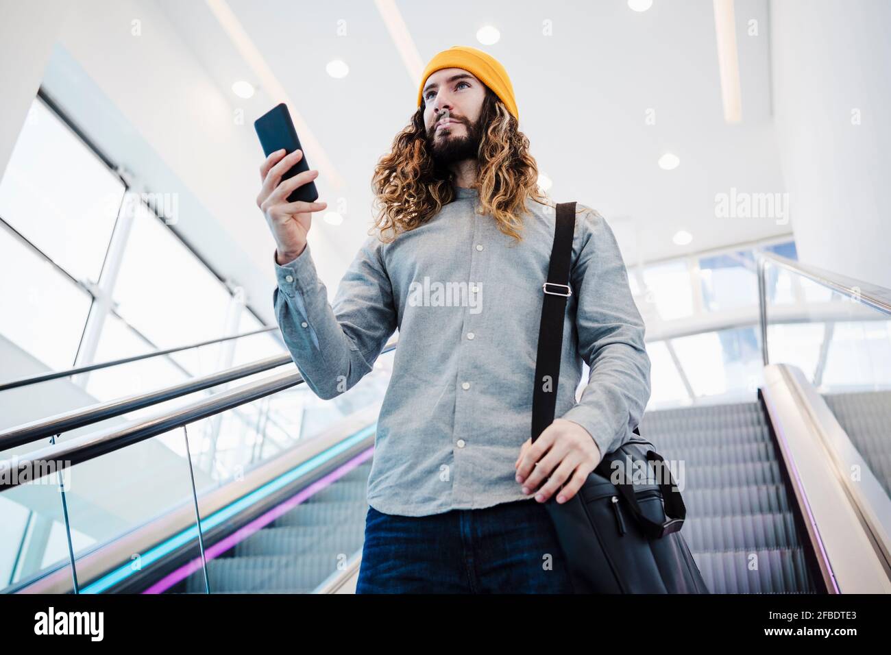 Homme avec de longs cheveux tenant le téléphone mobile tout en continuant escalier roulant Banque D'Images