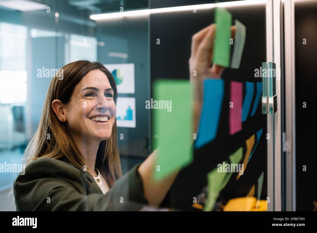 Une femme d'affaires excitée remue-méninges en utilisant des notes adhésives sur le mur de verre à bureau Banque D'Images