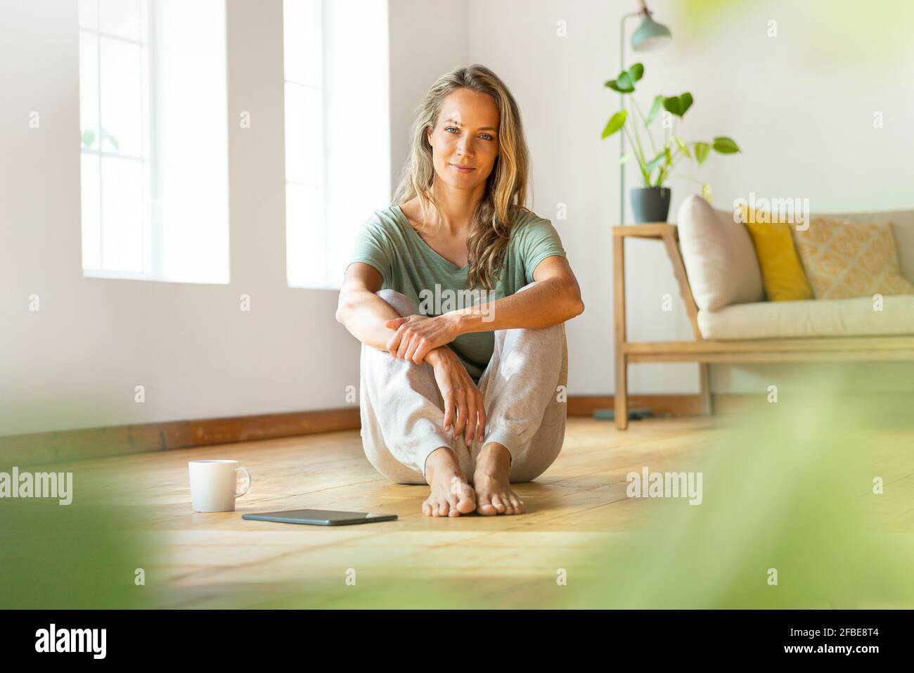 Femme confiante en vêtements de détente assise sur le sol par tablette numérique et tasse à café à la maison Banque D'Images