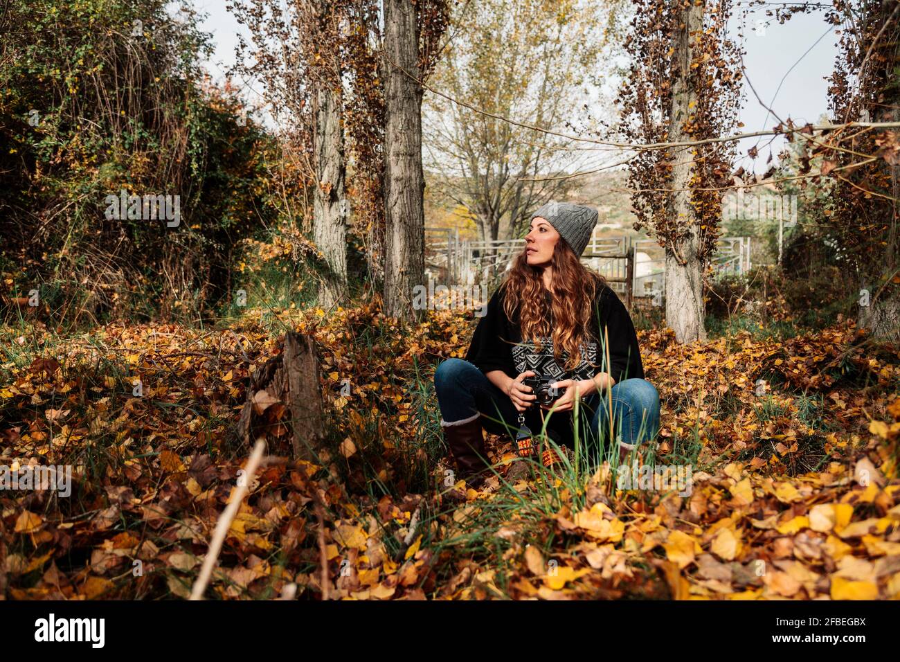 Femme avec un appareil photo qui regarde loin tout en étant assise au milieu des feuilles d'automne en forêt Banque D'Images