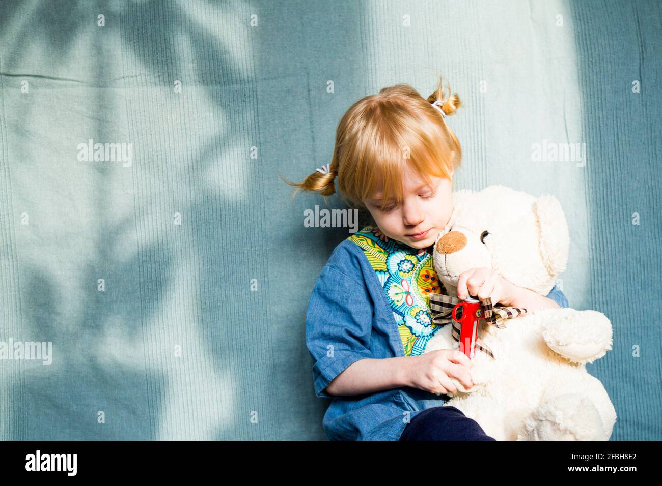 Fille blonde jouant avec l'ours en peluche contre le tissu bleu pendant jour ensoleillé Banque D'Images