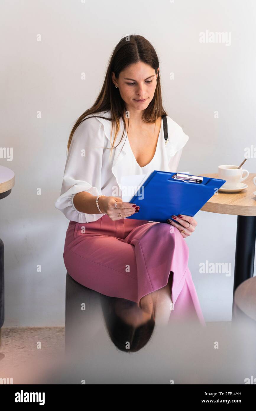 Jeune femme d'affaires vérifiant le fichier de presse-papiers lorsqu'elle est assise dans un café Banque D'Images