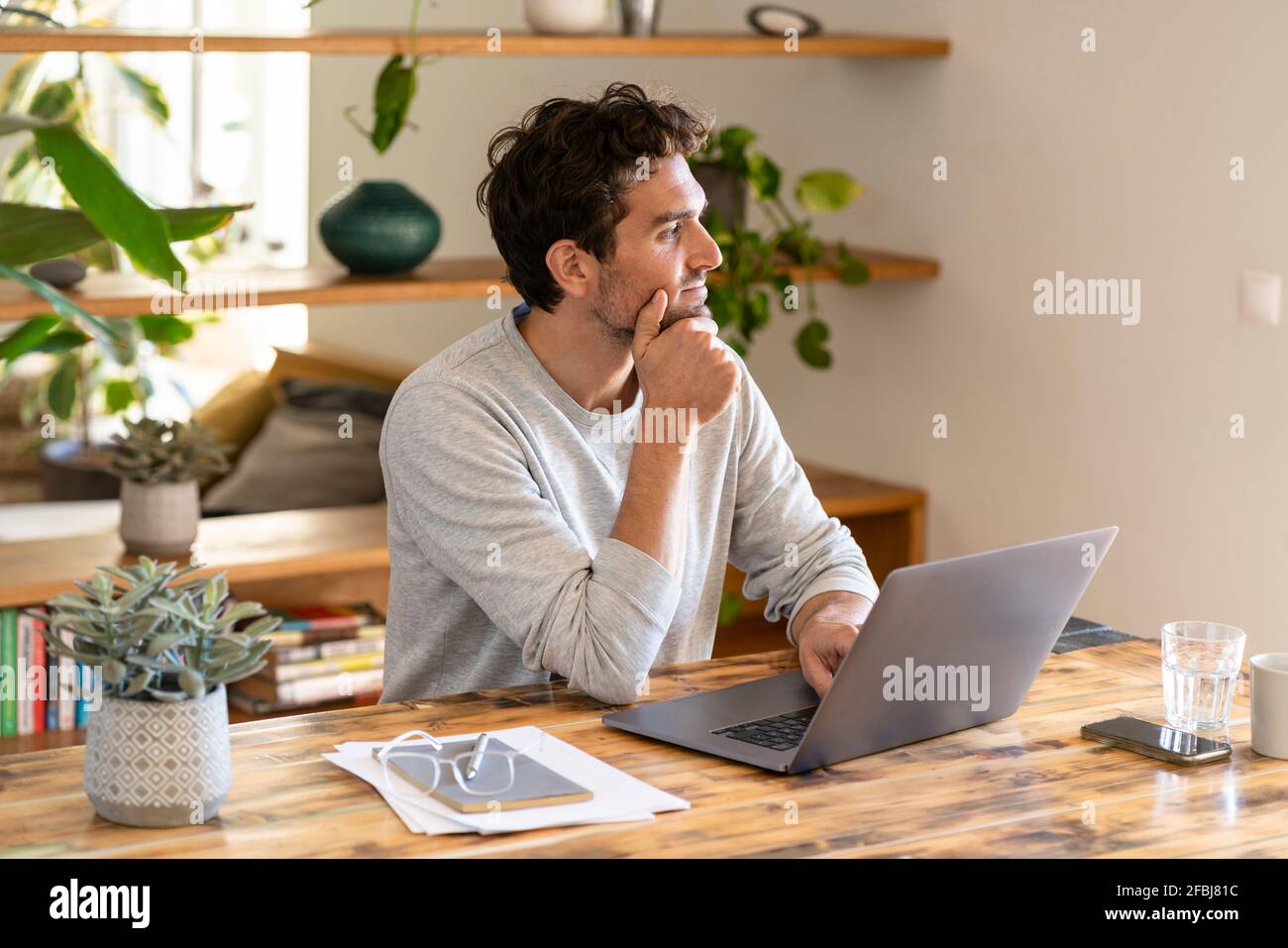 Contemplant le travailleur indépendant qui regarde loin tout en étant assis avec un ordinateur portable à bureau à domicile Banque D'Images