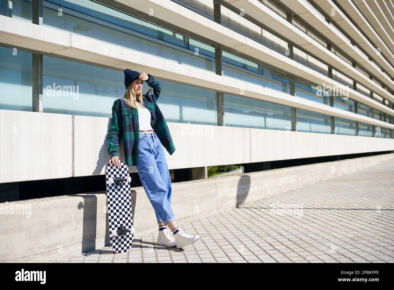 Jeune femme regardant loin en se tenant debout avec une planche à roulettes devant le bâtiment pendant la journée ensoleillée Banque D'Images Jeune femme regardant loin en se tenant debout avec une planche à roulettes devant le bâtiment pendant la journée ensoleillée Banque D'Images