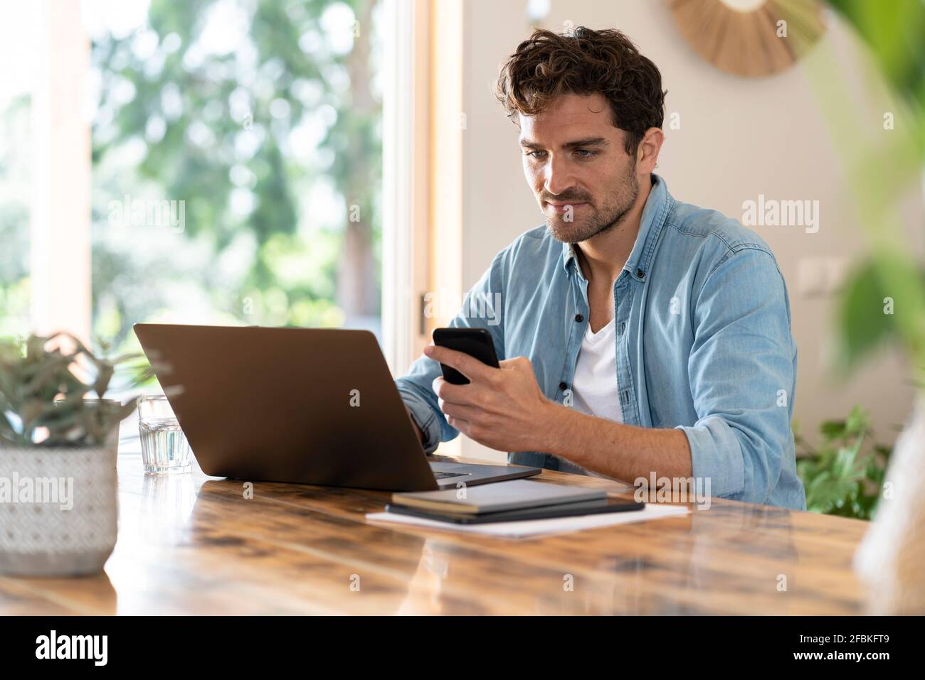 Travailleur indépendant sérieux tenant un téléphone portable tout en utilisant un ordinateur portable à la table Banque D'Images