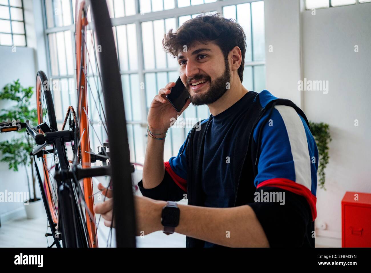 Jeune homme souriant parlant sur un smartphone tout en tenant un vélo rayons de roue à la maison Banque D'Images