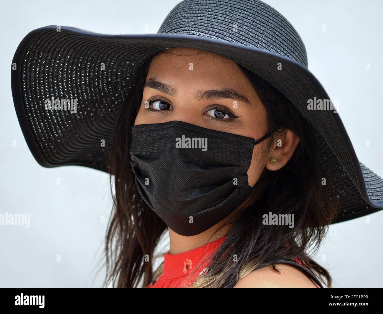 Une jeune femme brune mexicaine aux beaux yeux bruns porte un chapeau de soleil noir et un masque jetable noir pendant la pandémie du coronavirus. Banque D'Images