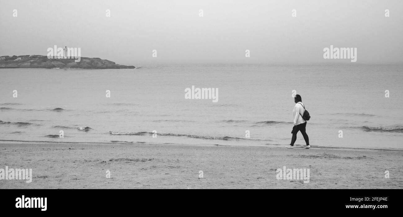 Image en noir et blanc d'une femme marchant pieds nus sur une plage floue, et d'un homme debout sur des rochers éloignés Banque D'Images