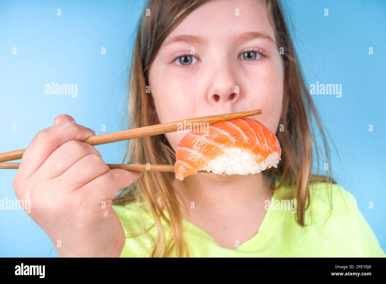 Une petite fille adorable aime manger des sushis. Joyeux drôle caucasien blonde petite fille avec divers rouleaux de sushi et baguettes. Sur fond bleu vif Banque D'Images