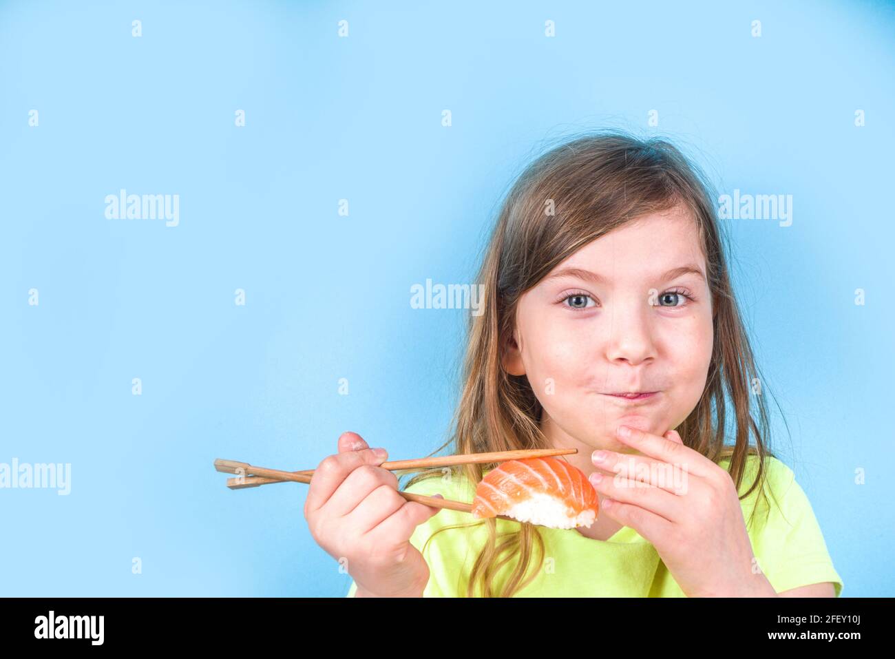 Une petite fille adorable aime manger des sushis. Joyeux drôle caucasien blonde petite fille avec divers rouleaux de sushi et baguettes. Sur fond bleu vif Banque D'Images