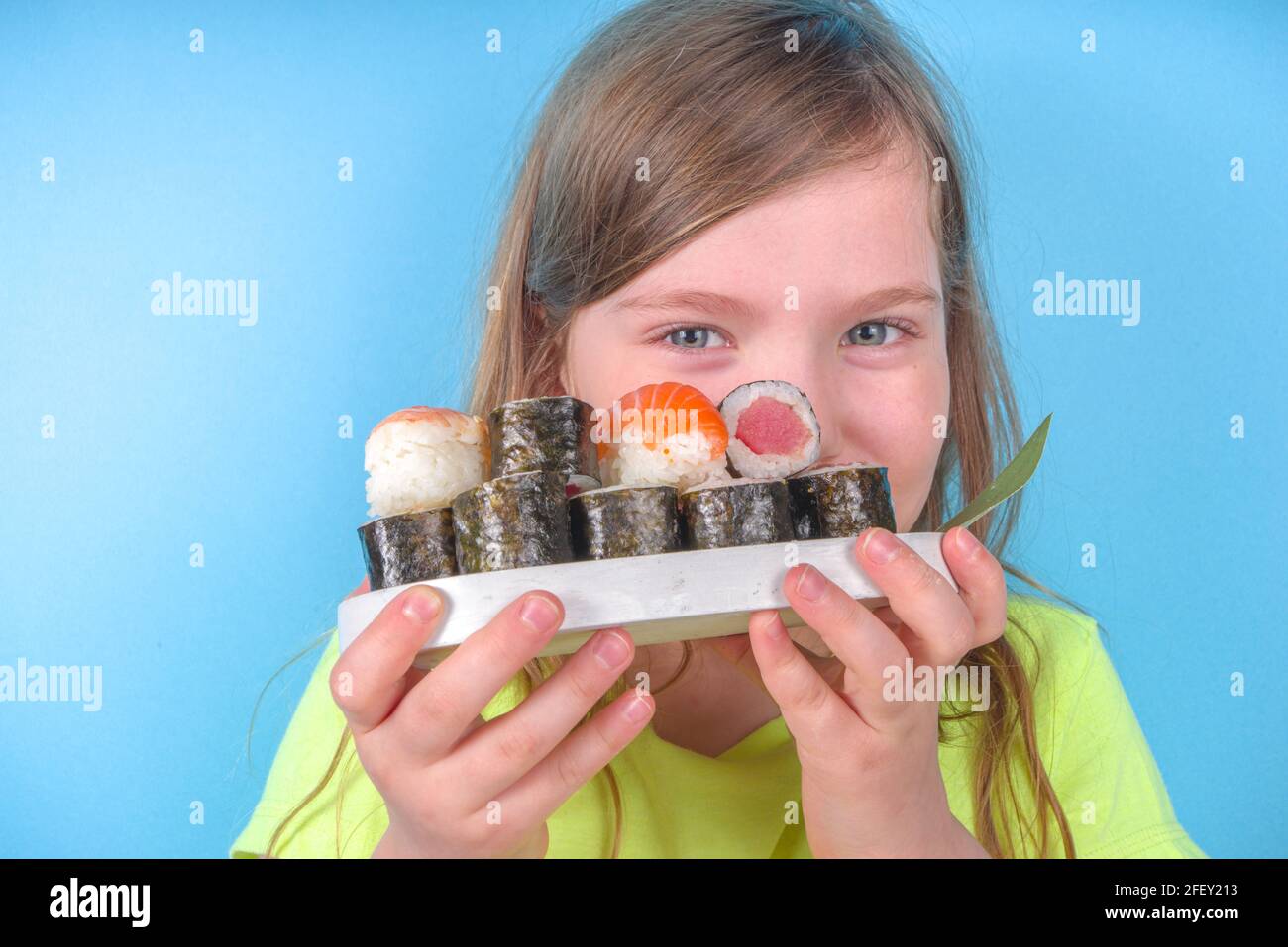 Une petite fille adorable aime manger des sushis. Joyeux drôle caucasien blonde petite fille avec divers rouleaux de sushi et baguettes. Sur fond bleu vif Banque D'Images
