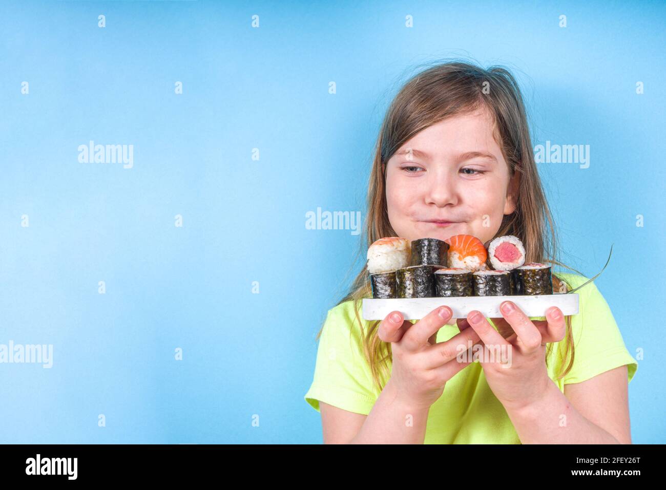 Une petite fille adorable aime manger des sushis. Joyeux drôle caucasien blonde petite fille avec divers rouleaux de sushi et baguettes. Sur fond bleu vif Banque D'Images