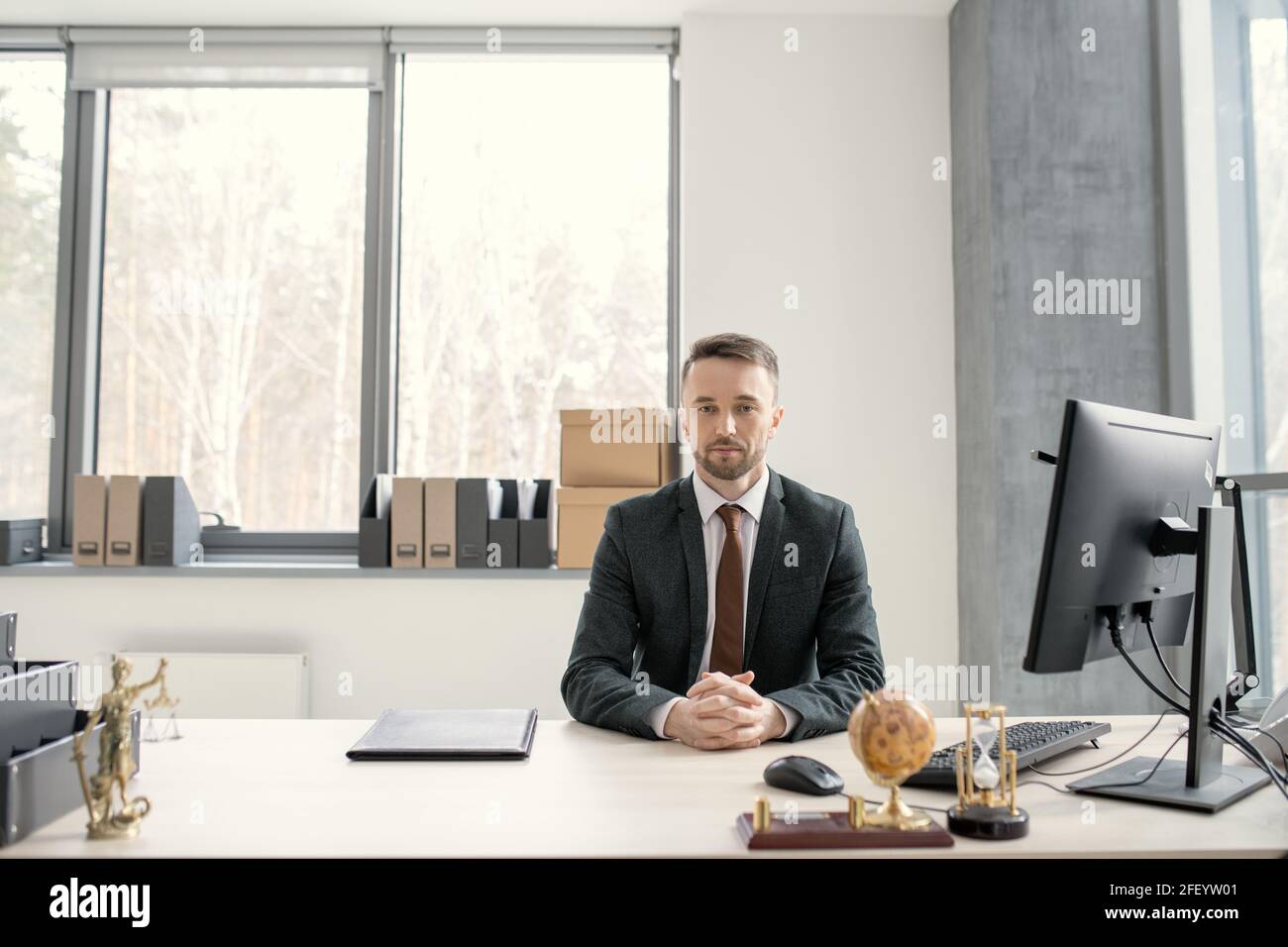 Portrait de sérieux beau jeune exécutif en costume noir et attachez-vous avec les mains clastées à la table équipée d'un ordinateur Banque D'Images