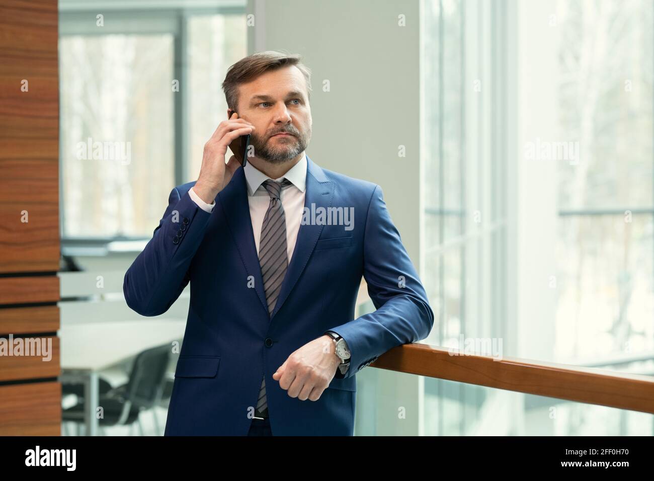 Homme d'affaires sérieux et attentionné d'âge moyen avec une barbe de trois jours penchée sur la main courante couloir de bureau et parler au téléphone Banque D'Images