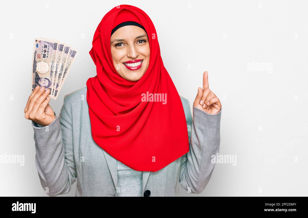 Belle femme hispanique portant le hijab islamique tenant des billets en yen japonais surpris par une idée ou une question pointant du doigt avec le visage heureux, engourdie Banque D'Images