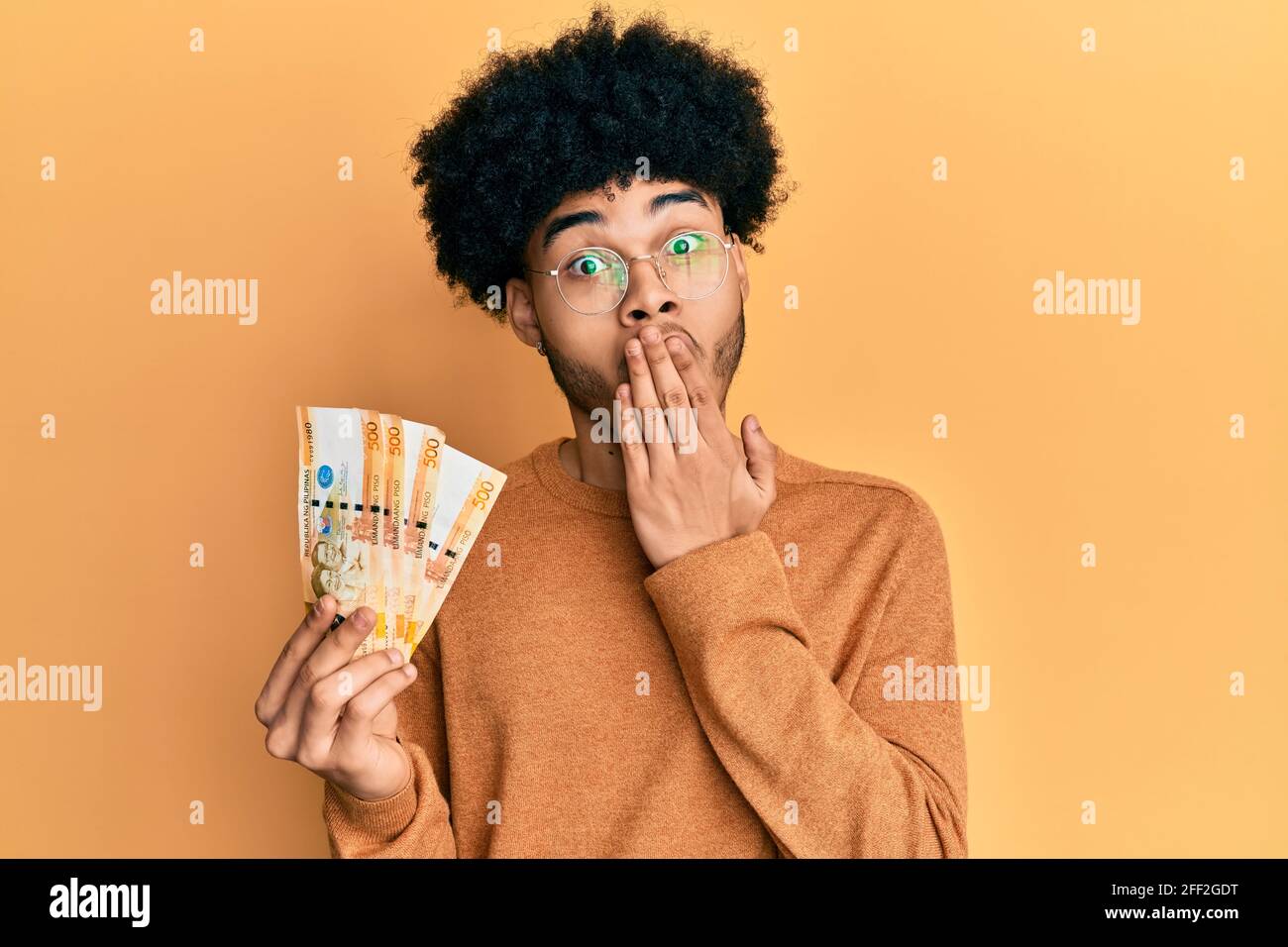 Jeune africain américain avec cheveux afro tenant 500 billets de banque de peso philippins couvrant la bouche avec la main, choqué et peur d'erreur. Surpris ex Banque D'Images