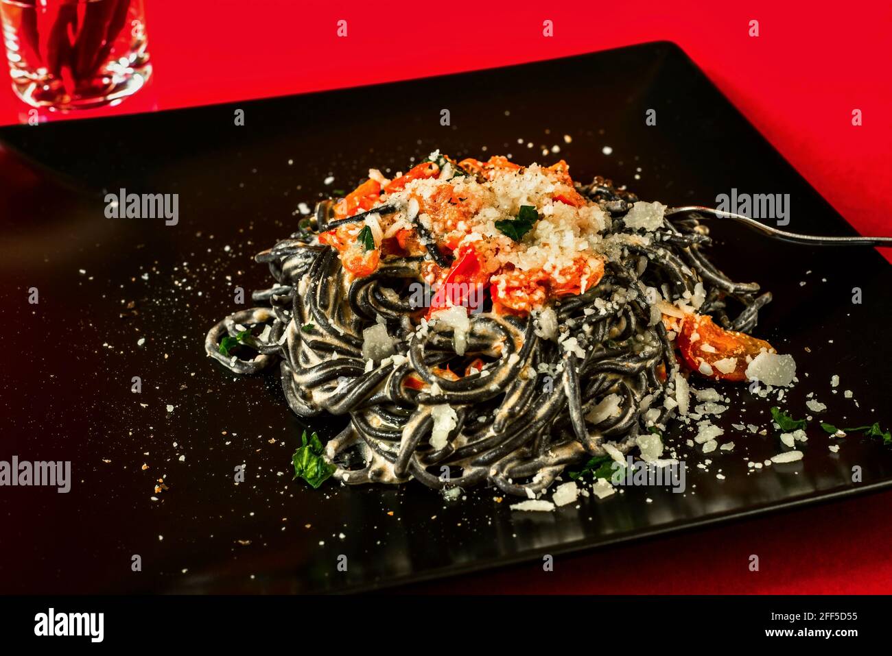 Spaghetti noirs avec tomate rôtie, piment, ricotta et fromage parmason sur une grande assiette noire sur fond rouge, gros plan. Banque D'Images