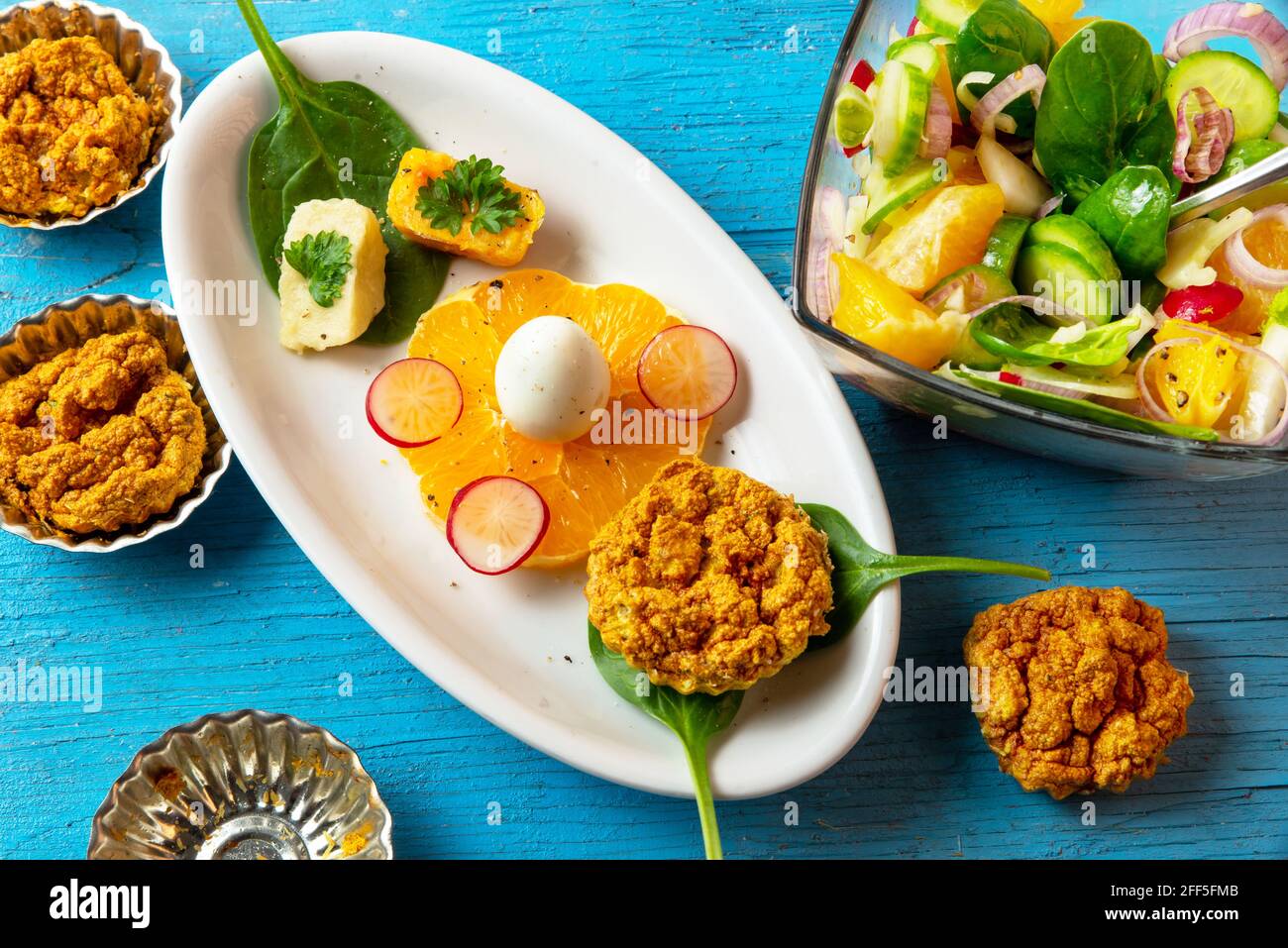 Table bleue avec assiette et bol avec salade de légumes et orange, cube avec purée de légumes et œuf de caille, petite pâtisserie arrondie et forme. Banque D'Images