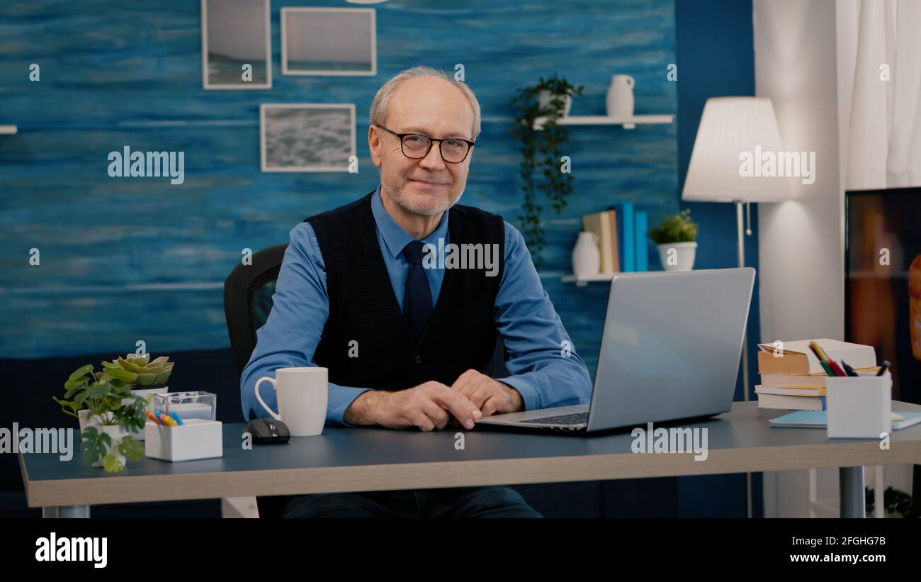 Homme senior regardant la caméra souriant assis sur le lieu de travail au bureau, travaillant de la maison tandis que la vieille femme regardait la télévision en arrière-plan. Personne d'affaires à la retraite préparée pour une réunion en ligne à l'aide de gadgets moden Banque D'Images